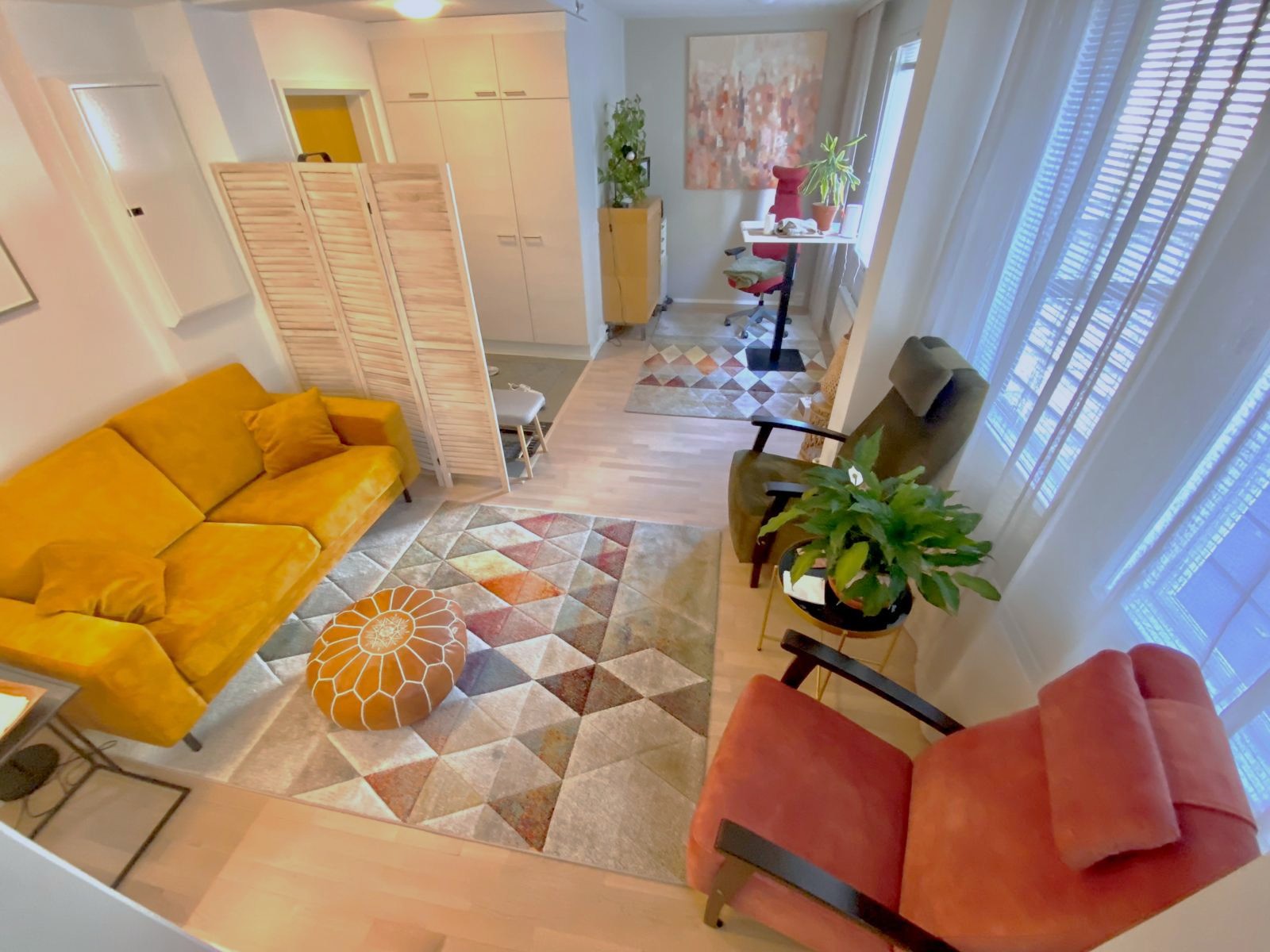Cozy room with a yellow sofa, orange armchair, and geometric-patterned rugs. Natural light filters through blinds.